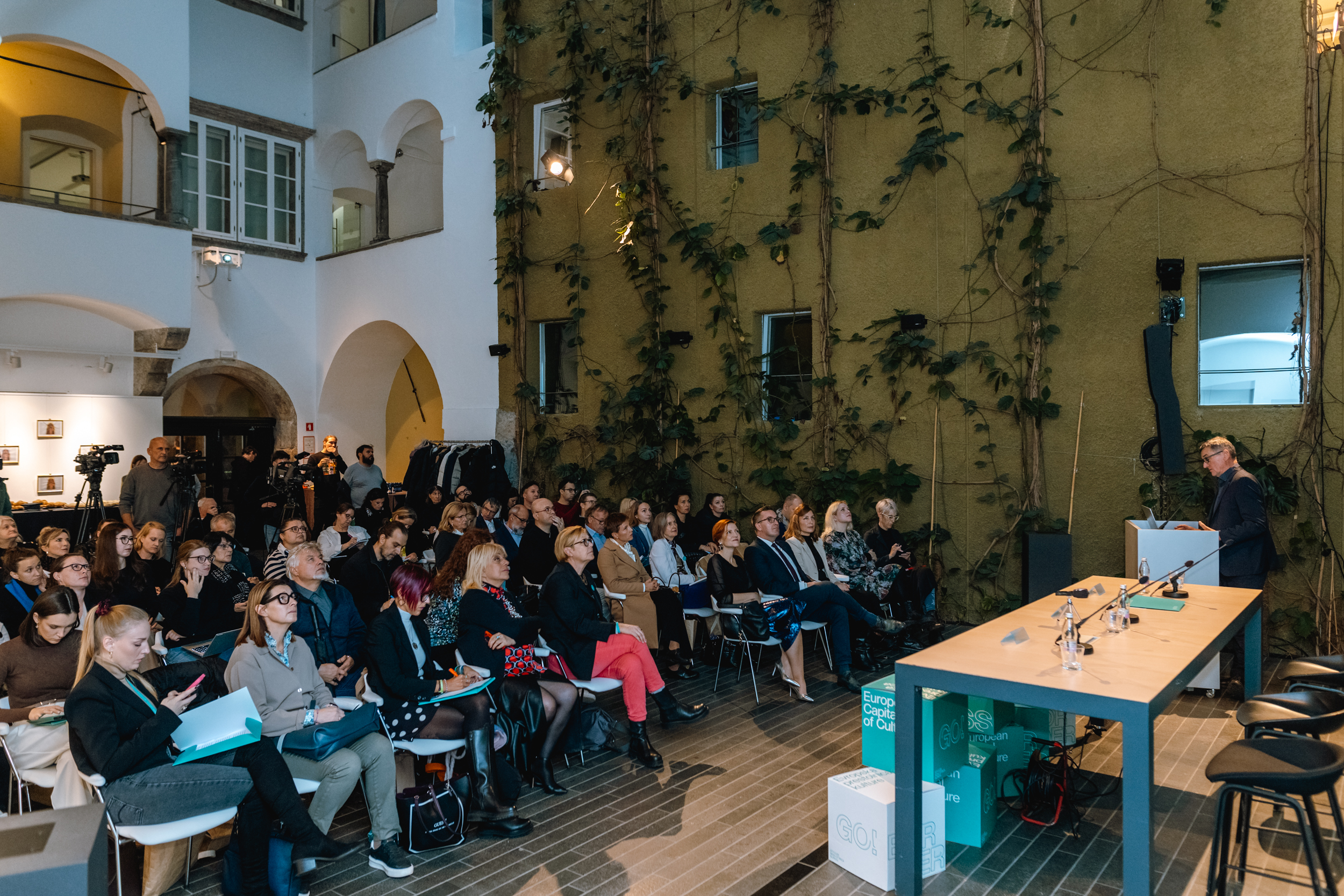 Veliko udeležencev sedi na stolih in spremlja novinarsko konferenco. Udeleženci sedijo v vrstah. Dogodek se dogaja v večernem času, saj je prostor osvetljen. Iz prostora je razvidno, da gre za zunanji avditorij. Na koncu prostora so kamere ter novinarji.