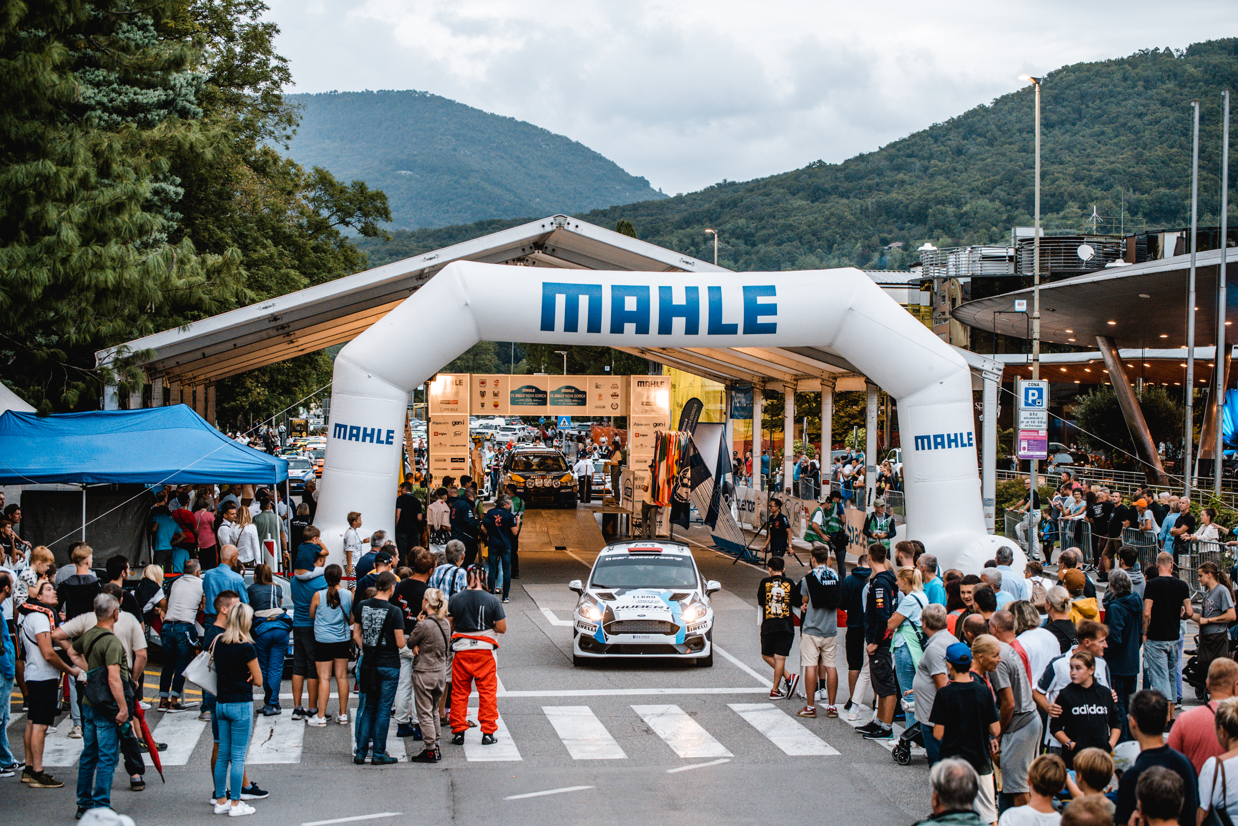 Dirkalni avto obkrožajo obiskovalci in gledalci rallyja. Dirkalni avto je bele avto z modrimi črtami. Na avtomobilu so zapisani sponzorji. Avto stoji pred napihljivim belim lokom, na katerem piše MAHLE. Za avtomobilom je velik šotor.