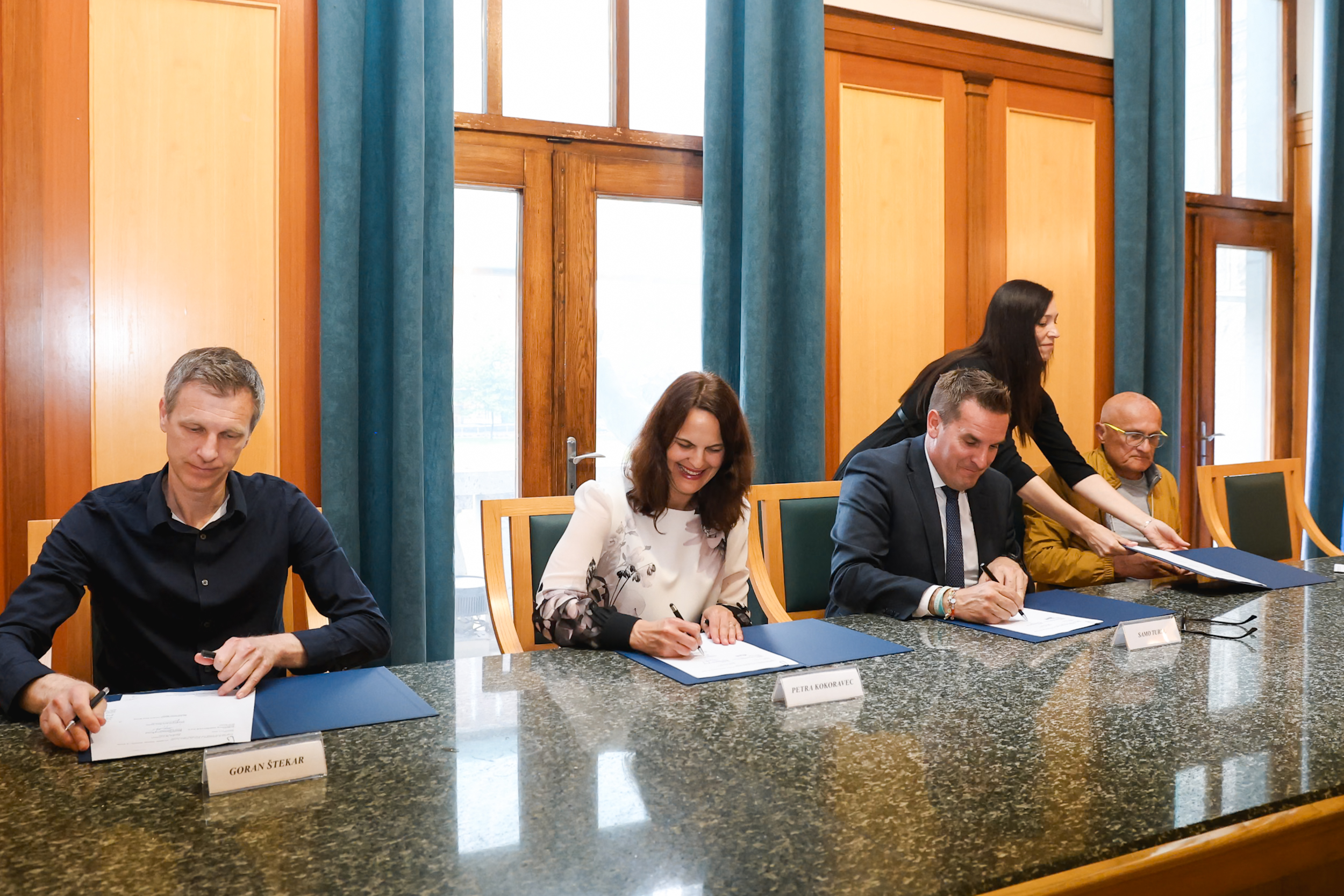 Z leve proti desni. Goran Štekar, Petra Kokoravec, Samo Turel, Miran Mihalič podpisujejo pogodbo. Osebe na sliki podpisujejo pogodbo z nasmeškom na obrazu.