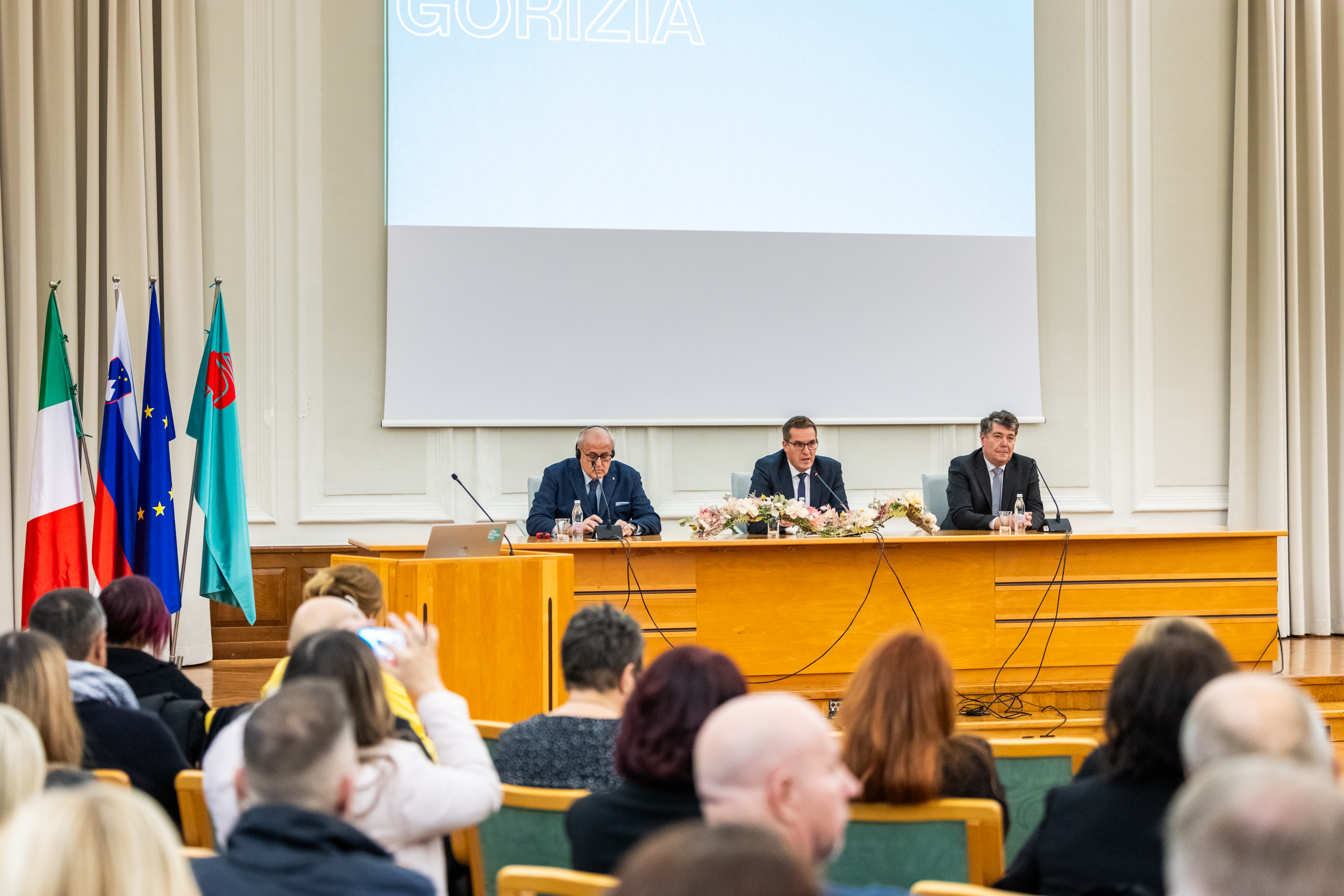 Trije župani sedijo za govorniškim pultom, pred njimi na stolih sedijo občinski svetniki. Za njimi je postavljeno veliko platno. Na njihovi levi strani so postavljene slovenska, italijanska in evropska zastava ter zastava Mestne občine Nova Gorica.