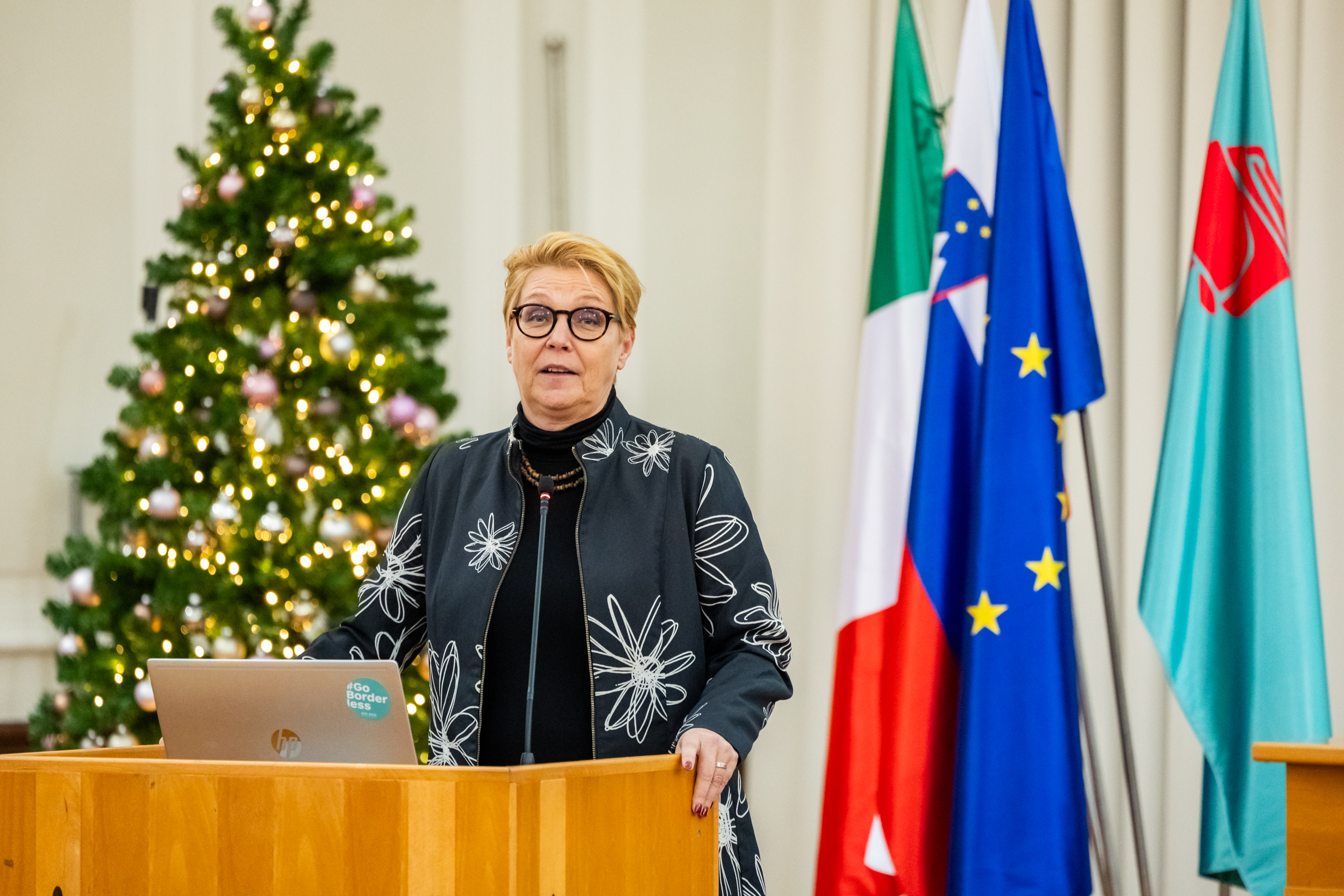 Režiserka otvoritvene slovesnosti Neda Rusjan Bric podaja svoj nagovor za govorniškim pultom. Pred njo je postavljen prenosni računalnik z mikrofonom. Za njo se vidi okrašeno novoletno drevo ter slovenska, italijanska in evropska zastava ter zastava Mestne občine Nova Gorica.