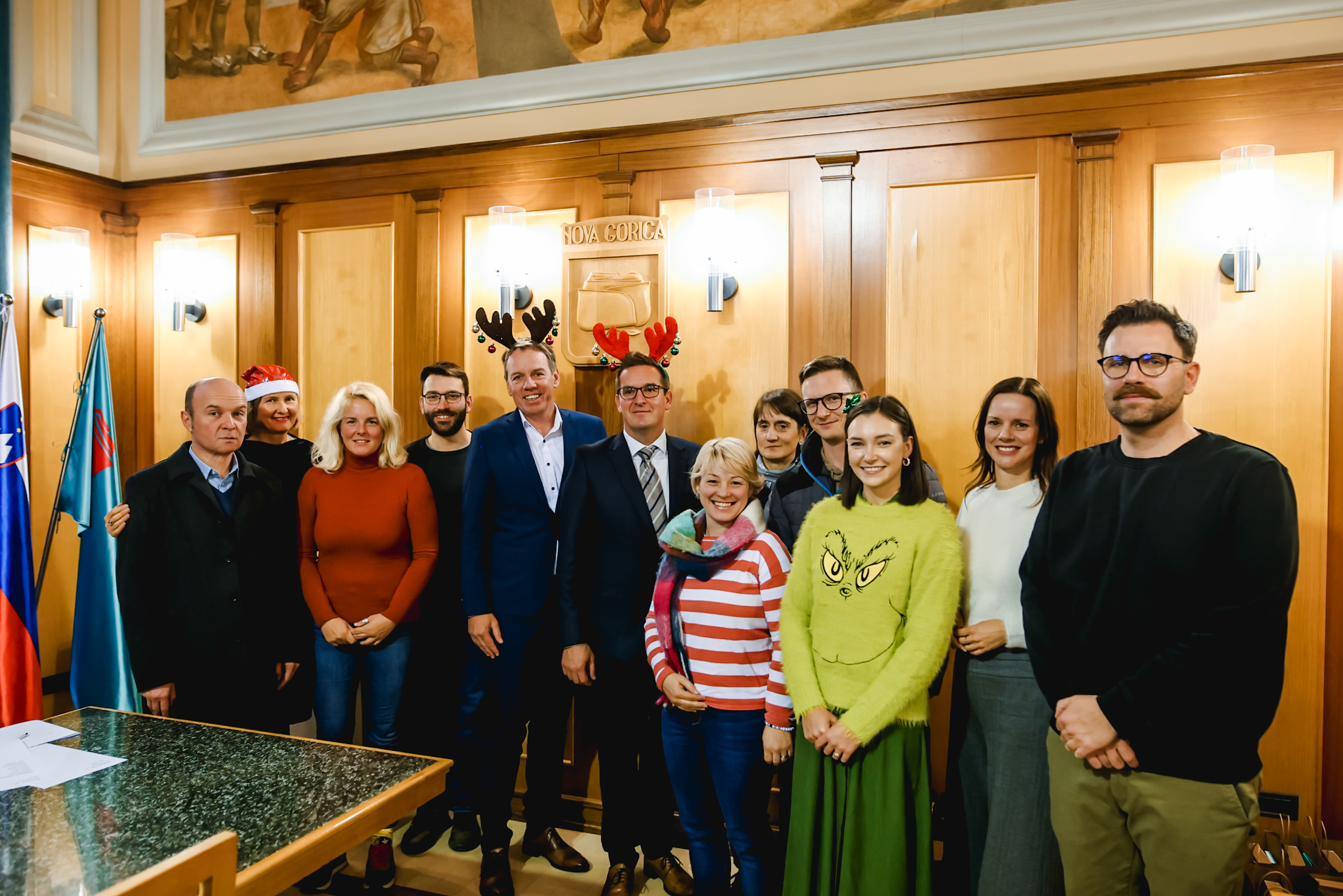 Skupinska fotografija dvanajstih nasmejanih udeležencev novinarske konference, ki skupaj stojijo ob steni v Zeleni dvorani novogoriške Občinske palače.